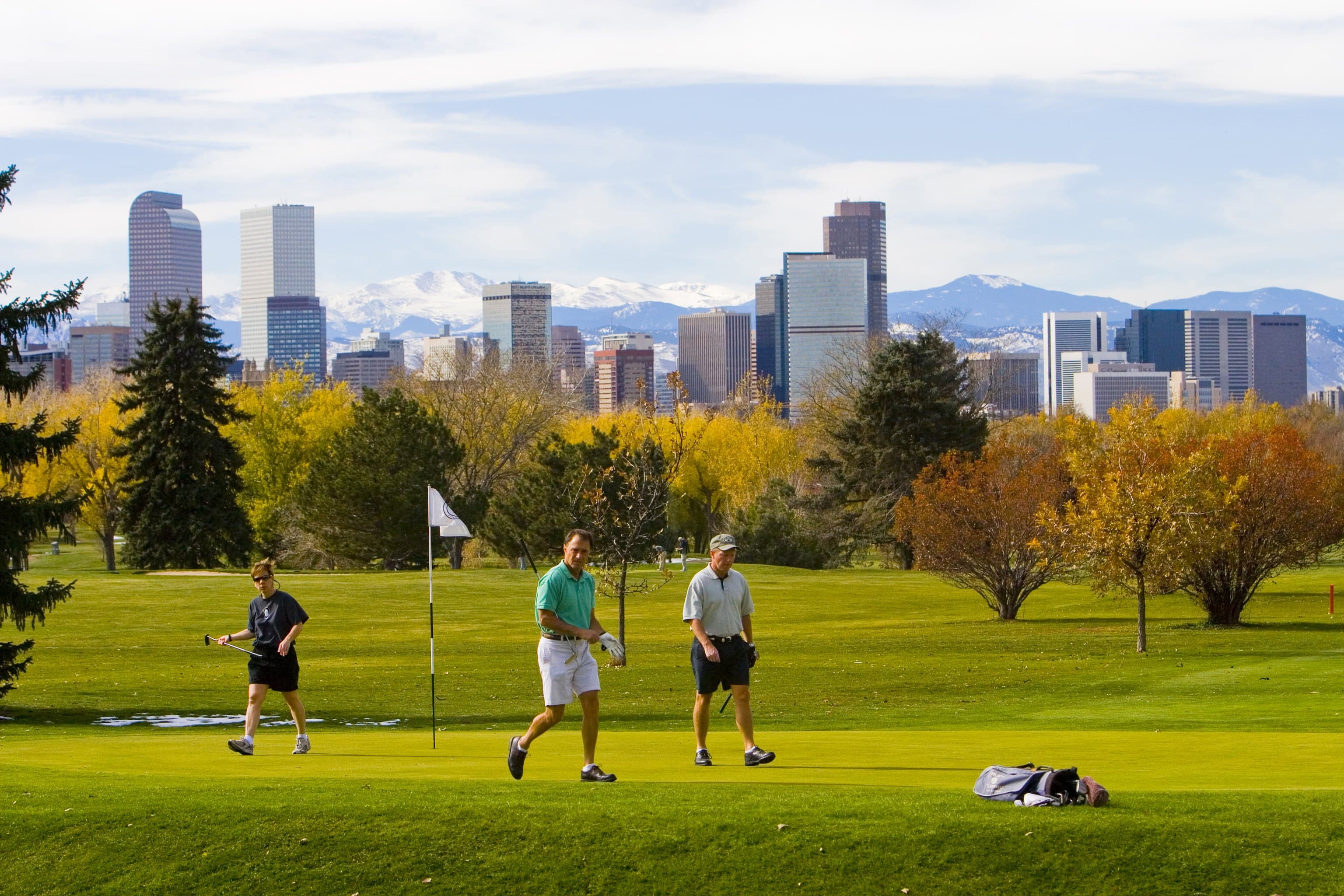 Three men in summer gear walk the green fairways of the golf course. From this view, we can see the many skycrapers of downtown Denver and snowcapped peaks farther west.