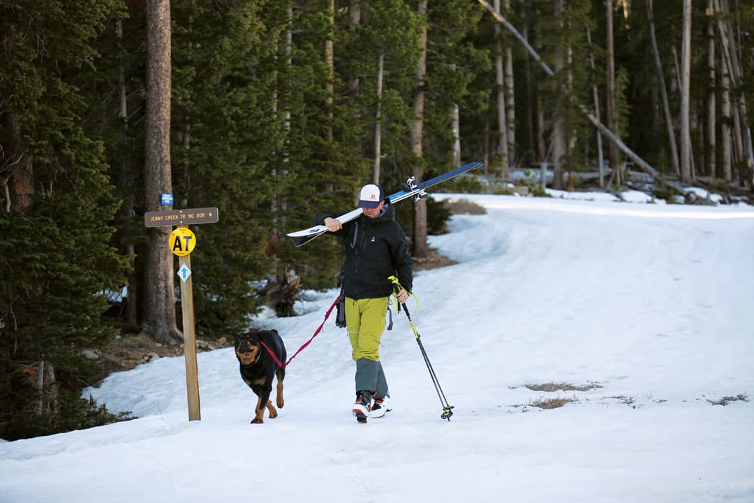 A man with carrying skis across his shoulder walks down a snowy path with a dog on a leash