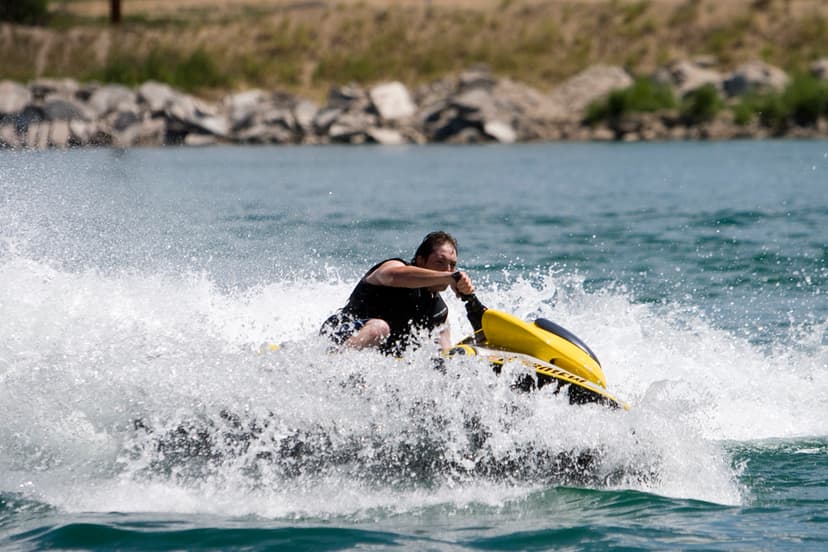 A person sits on a yellow jet ski in a large white wave on Jackson Lake. On the shore behind them there are big rocks and boulders that meet greenery.