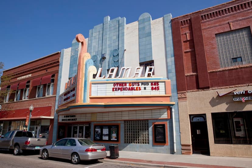 The Art Deco Lamar Theater sandwiched in between red-brick buildings. The theater is painted white, blue and orange. The marquee says "Other Guys PG13 545 Expendables R 645."