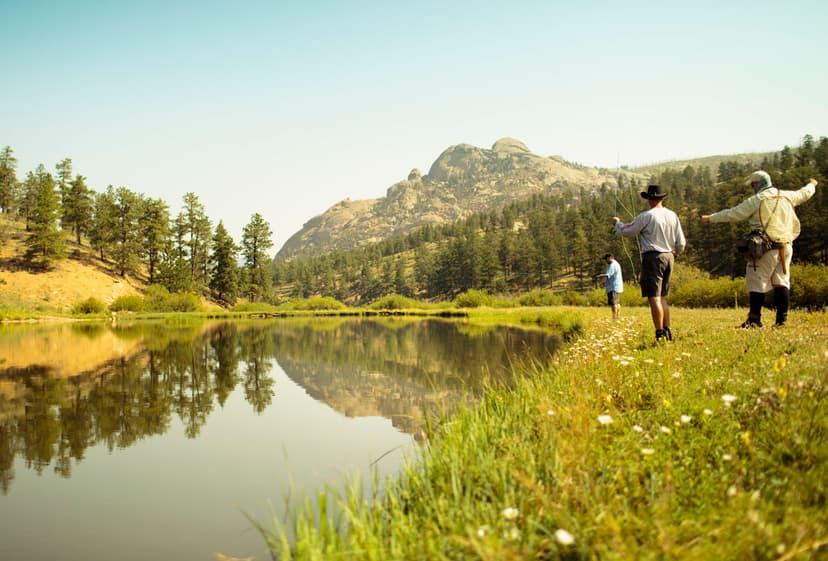 Three people fish on the green-yellow, grass-covered riverbank. The river is glassy, reflecting the evergreen trees and mountain in the background. The light-blue sky is hazy and the image has a yellow hue.