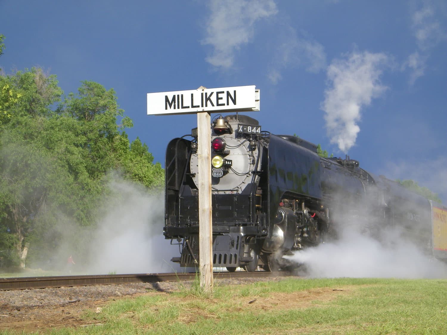 A historical black train engine creates steam on a track that is next to grass and green-leafed trees. In the middle of the image a white rectangular sign says "Milliken" in black letters.