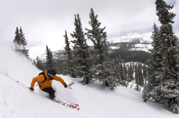 A skier wearing a yellow jacket skis down a snowy slope with a view of snow-covered mountain peaks in the distance.
