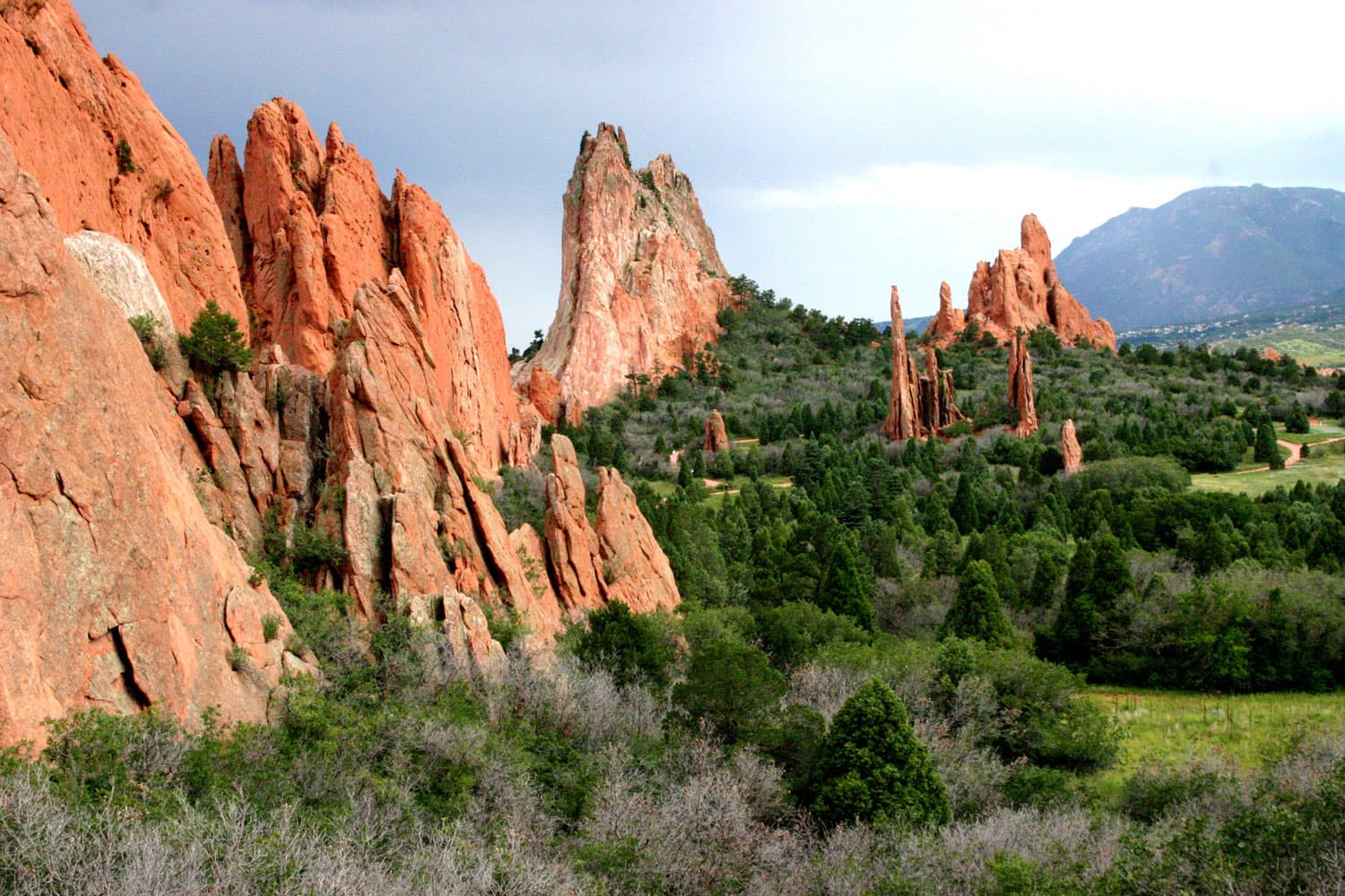 On the left side, thin red-rock formations protrude from the earth. On the right green-grass fields lead up to dark-green bushes at the base of the geologic formations at Garden of the Gods.