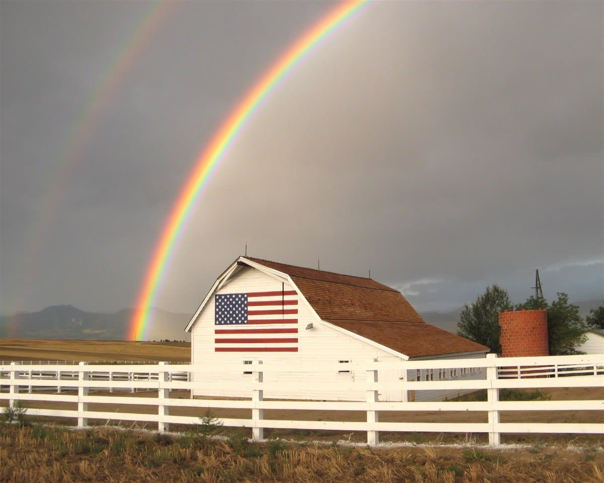 A rainbow protrudes from the grey, stormy sky above a white barn with an American flag painted on it.