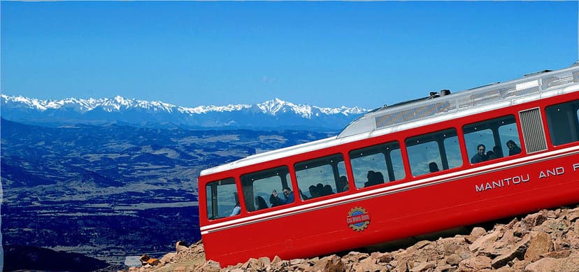 A bright red passenger compartment on a new train summits the rocky mountainside of America's Mountain in Colorado. Snow-capped peaks appear in the distance.