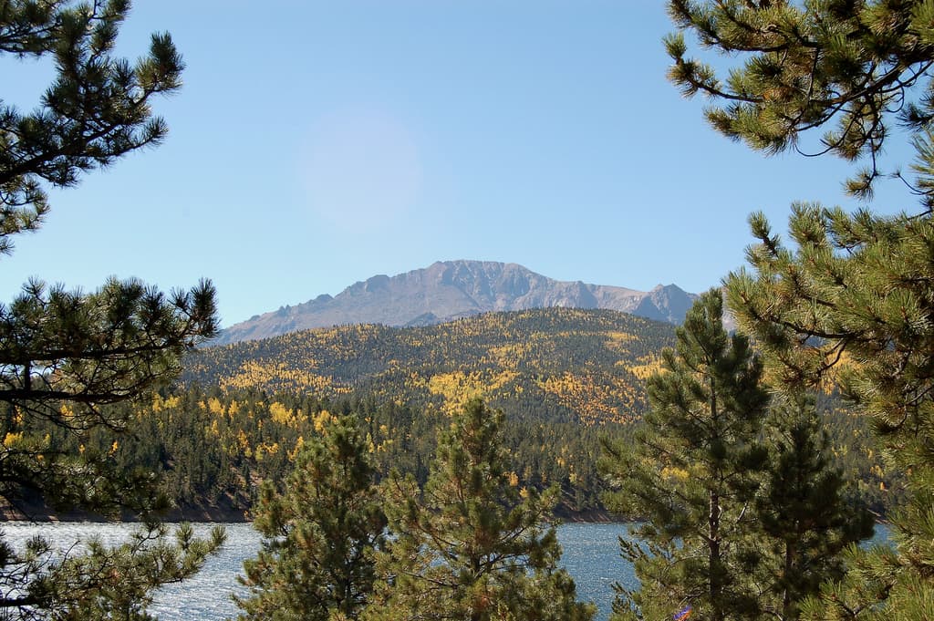Pikes Peak sits beneath a blue sky with a sparkling lake in the foreground. There are evergreen trees surrounding both sides of the frame and golden leaves are just starting to appear on trees.