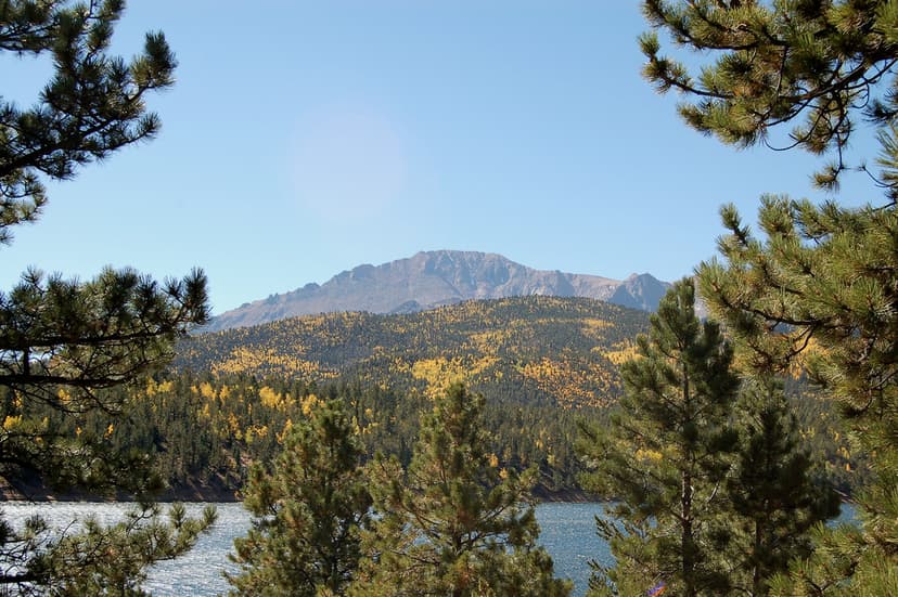 Pikes Peak sits beneath a blue sky with a sparkling lake in the foreground. There are evergreen trees surrounding both sides of the frame and golden leaves are just starting to appear on trees.