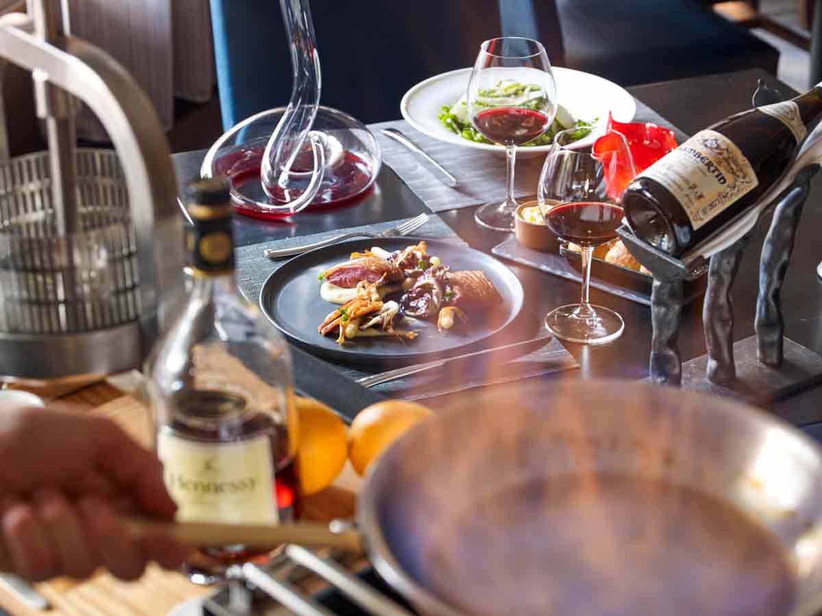 Two stemmed wine glasses sit on a table with plates of food and bottles of wine and spirits at The Little Nell in Aspen, Colorado.
