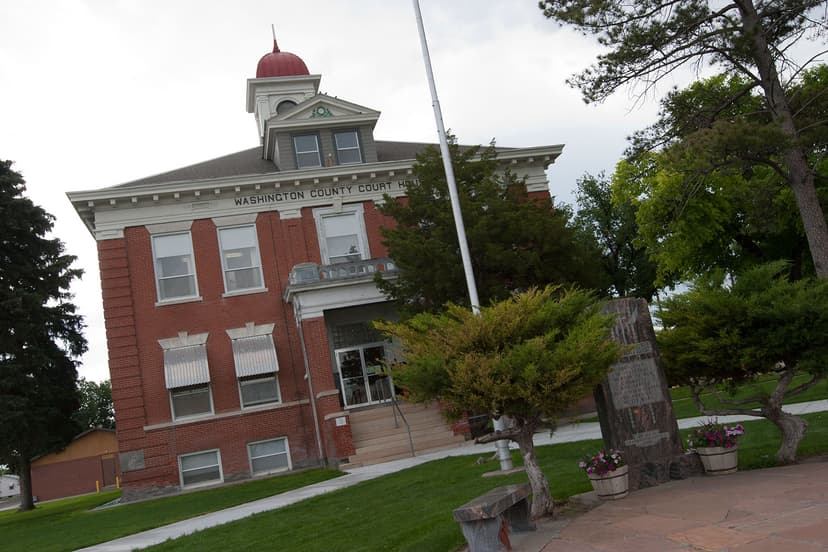 Front of the red-brick, two story Washington County Courthouse in downtown Akron, Colorado. The trees and grass are green. The cupola at the top of the building is red. 