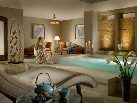 A person in a white robe sits on the raised ledge of a marble soaking pool at The Spa at the Arabelle hotel in Vail, Colorado. The room is decorated with golden twisting sculptures, pillar candles and chaise lounges.