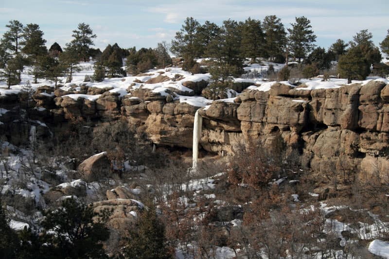 In the middle, a brown rock ridge is covered in snow with a ice waterfall cascading down. In the foreground, leafless trees sit with snow on the ground. On top of the ridge, evergreen trees.