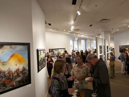 A crowd mingles in an art gallery with paintings lining the walls. 