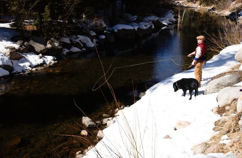 A man fly fishes on a snowy riverbank with his black dog on a river near Vail. Across the water, the riverbank is covered in boulders.