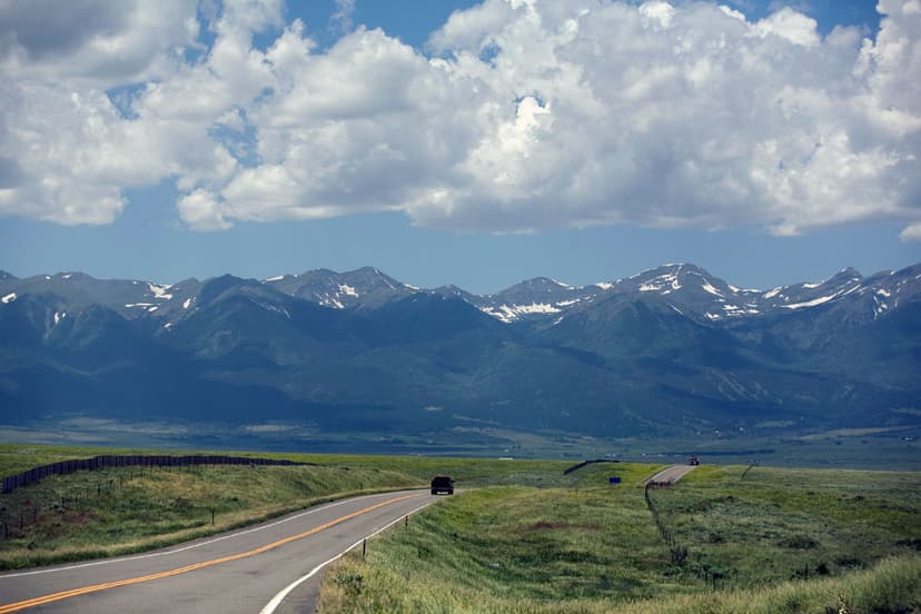 A grey highway with yellow stripes down the middle leads to snow-capped mountains. On both sides of the road are green farmlands.