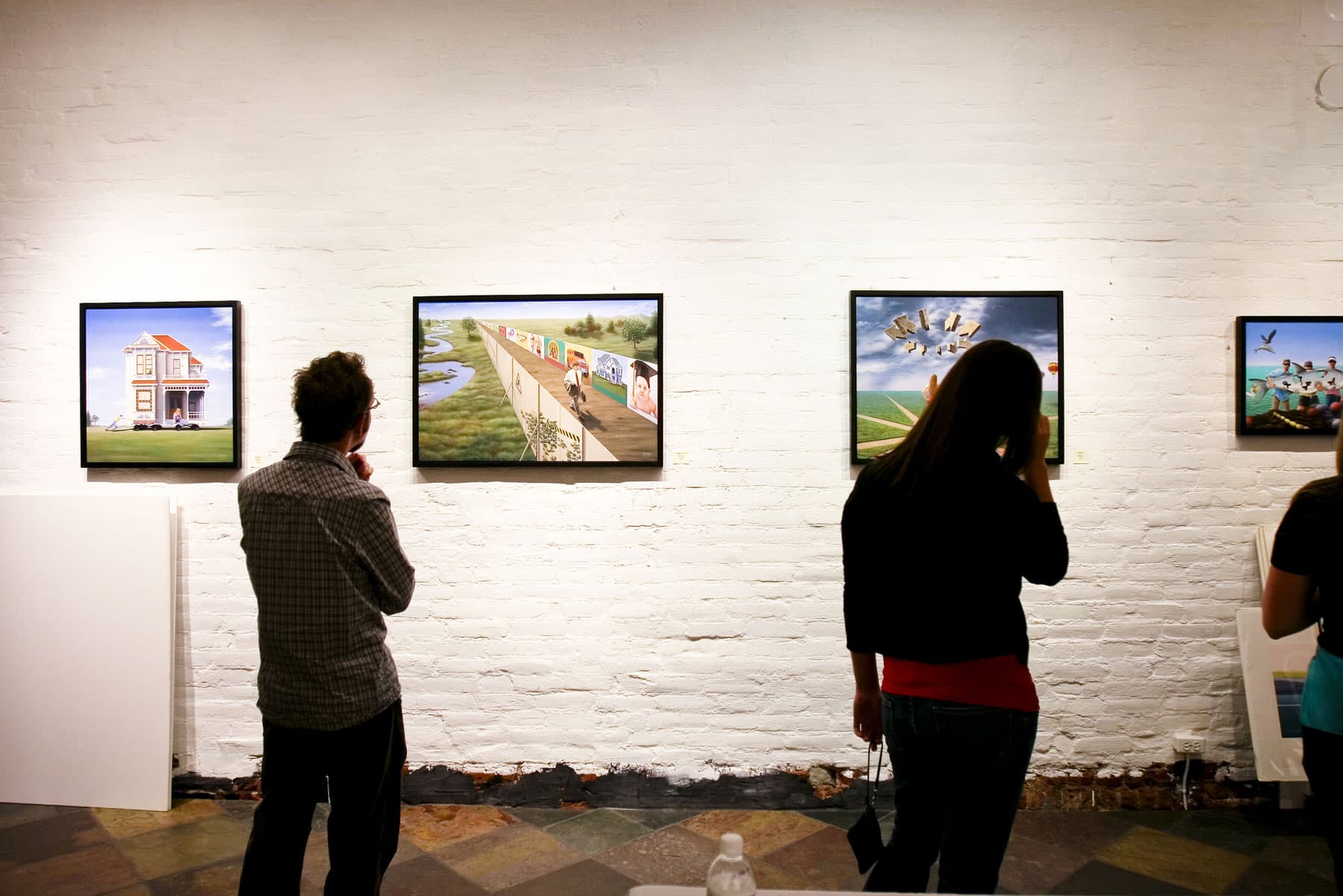 Four colorful painting with simple, black frames are evenly spread out across a white-painted brick wall. Attendees of Denver's Denver Arts Week admired the images on the wall.