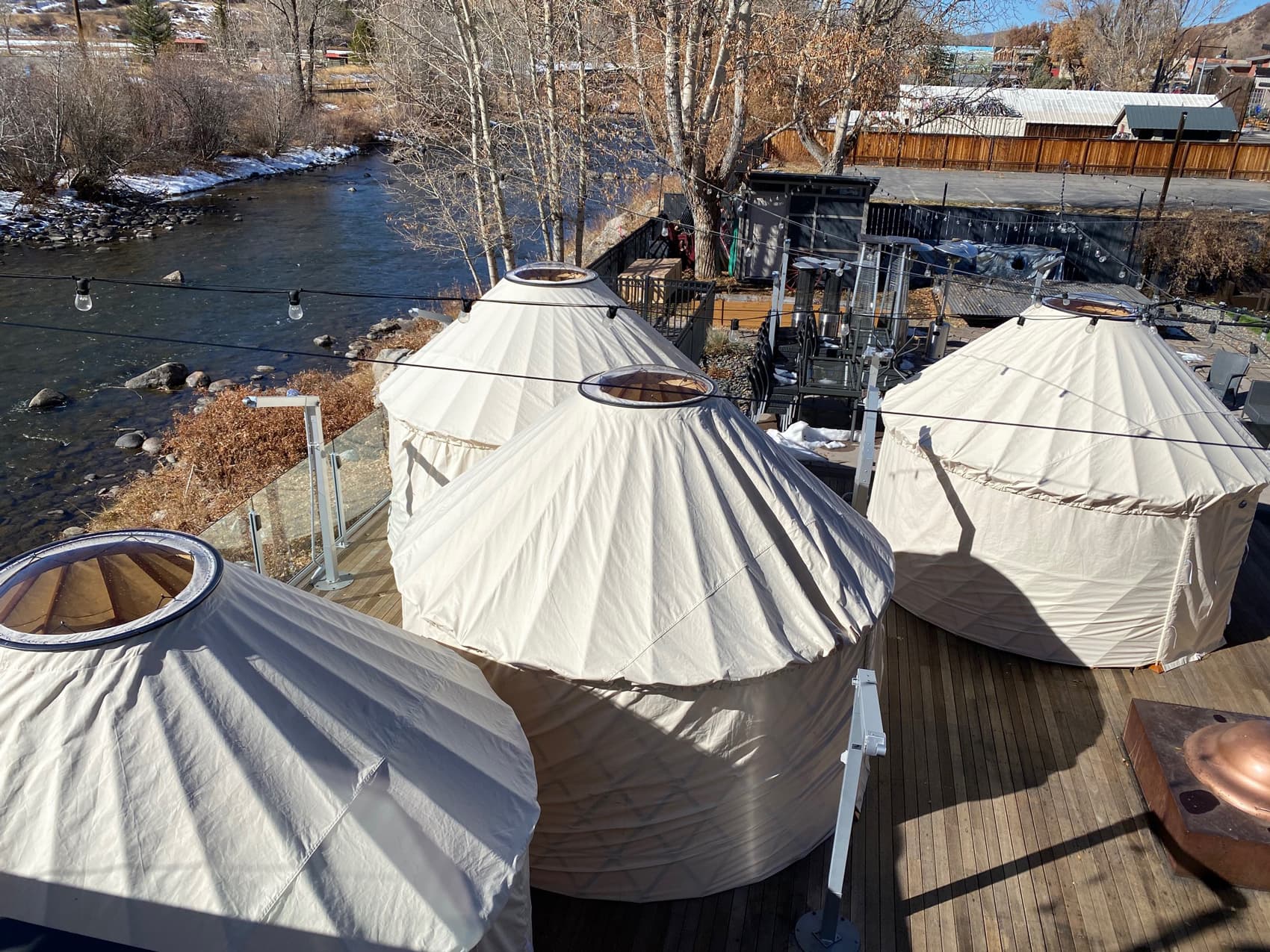 Four off-white yurts with holes at the top of the tents are set up at an event space alongside the flowing body of water in Steamboat Springs in winter.