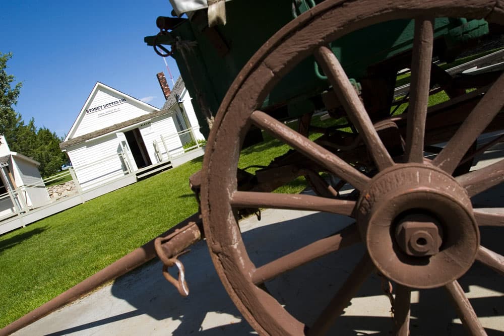A close up, tilted image of a brown wagon wheel with a white wooden building with a pediment in the background.