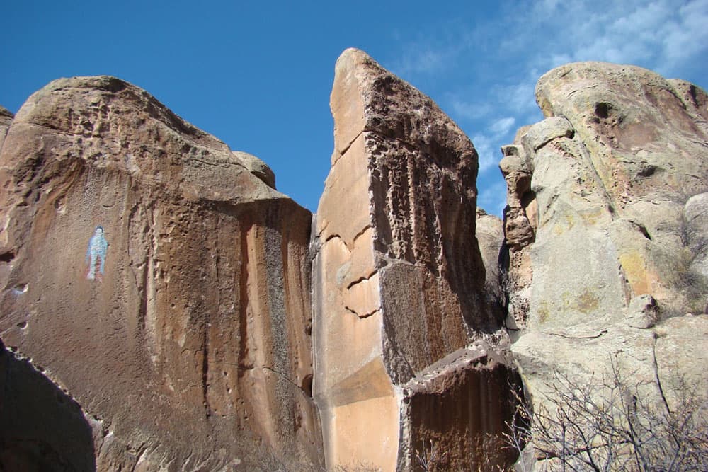 A tall, wall-like rock formation features a brightly painted human figure at Penitente Canyon in Colorado. A person scales the side of the canyon.