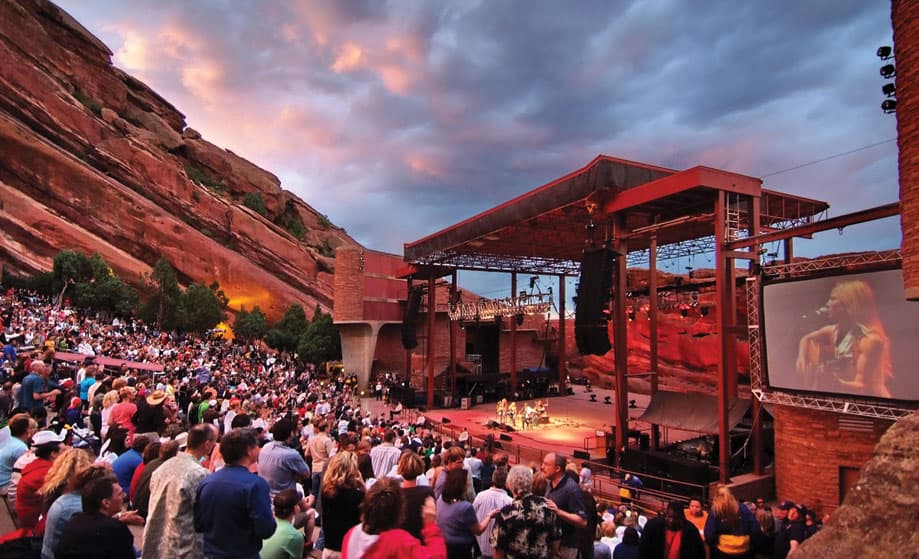 A crowd enjoys a musical performance at the Red Rocks Amphitheatre in Morrison, Colorado. A jumbo screen on the side shows a performer playing guitar.