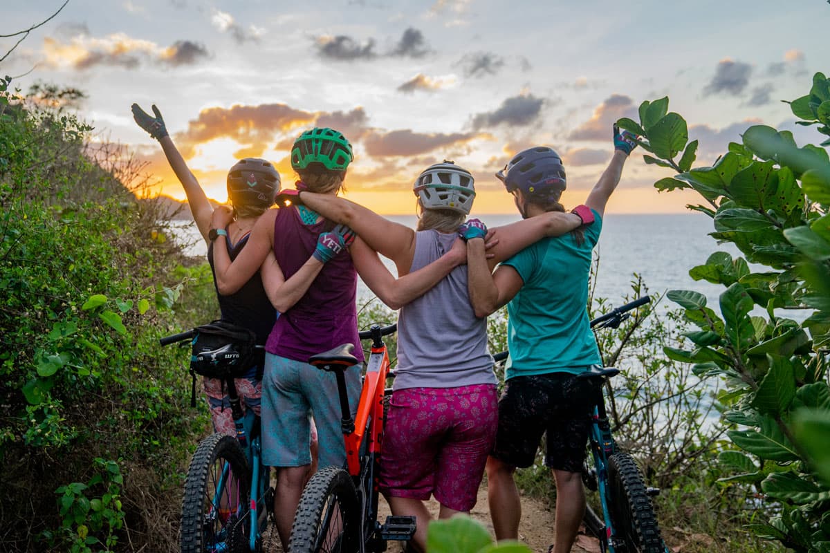 Four cyclists wearing outdoor gear drape their arms over each other or raise their arms to the run as they lookout over a lake in Colorado.