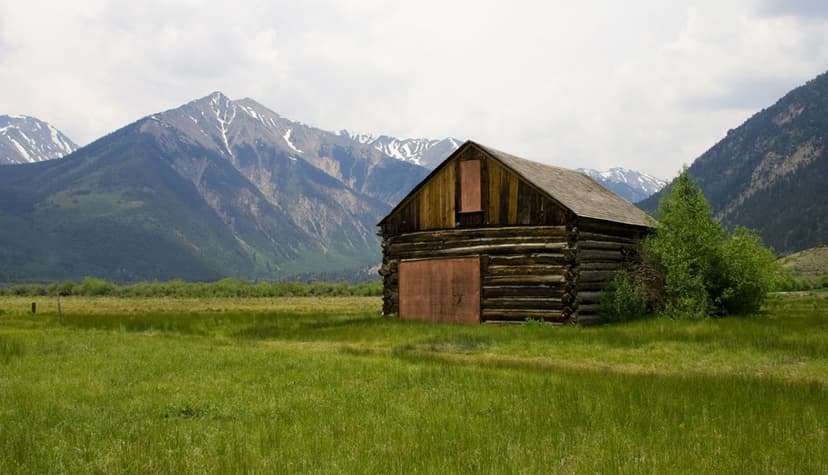 An old, wood cabin sits in a green field with hazy grey-topped mountains in the background with snow rivulets on Top of the Rockies Scenic Byway.