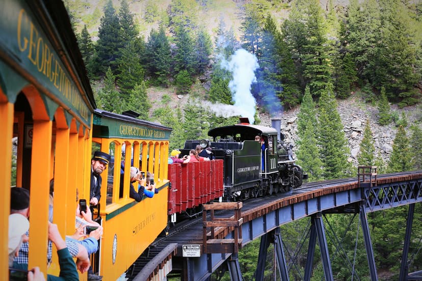 Smoke billows out of a train with yellow and red train cars. People are looking out the open windows as the cross a bridge over a valley with green trees in the background