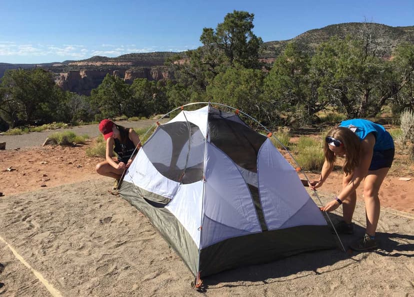 Two people setting up a small white tent at a campsite with trees in the background