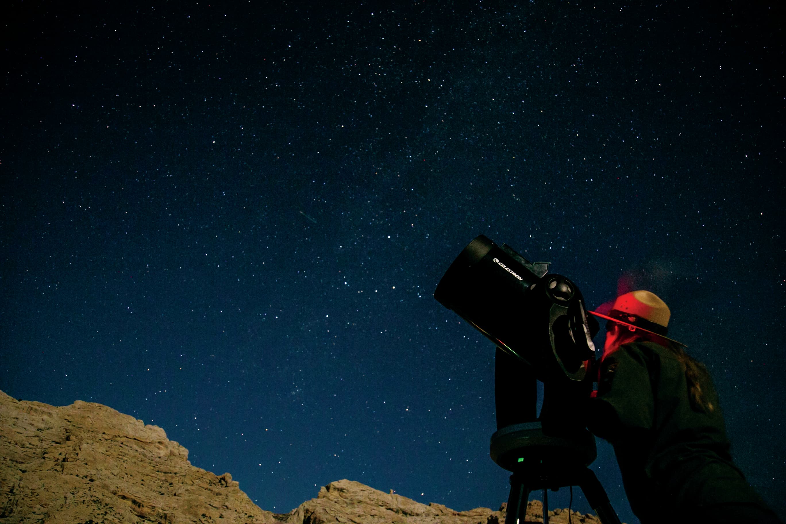 Someone in a hat looking into a telescope that's pointed at a star-studded sky.