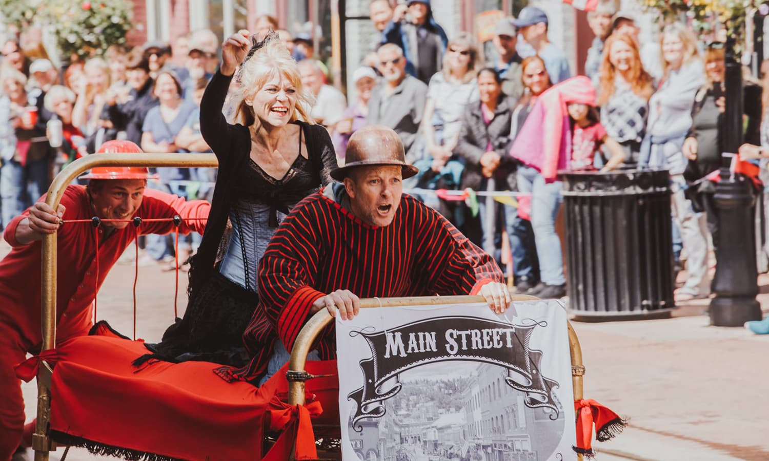 Madam Lou Bunch Bed Race with man in hat holding onto footboard, woman dressed in black behind him on the bed and man in red pushing the bed down the Main Street.