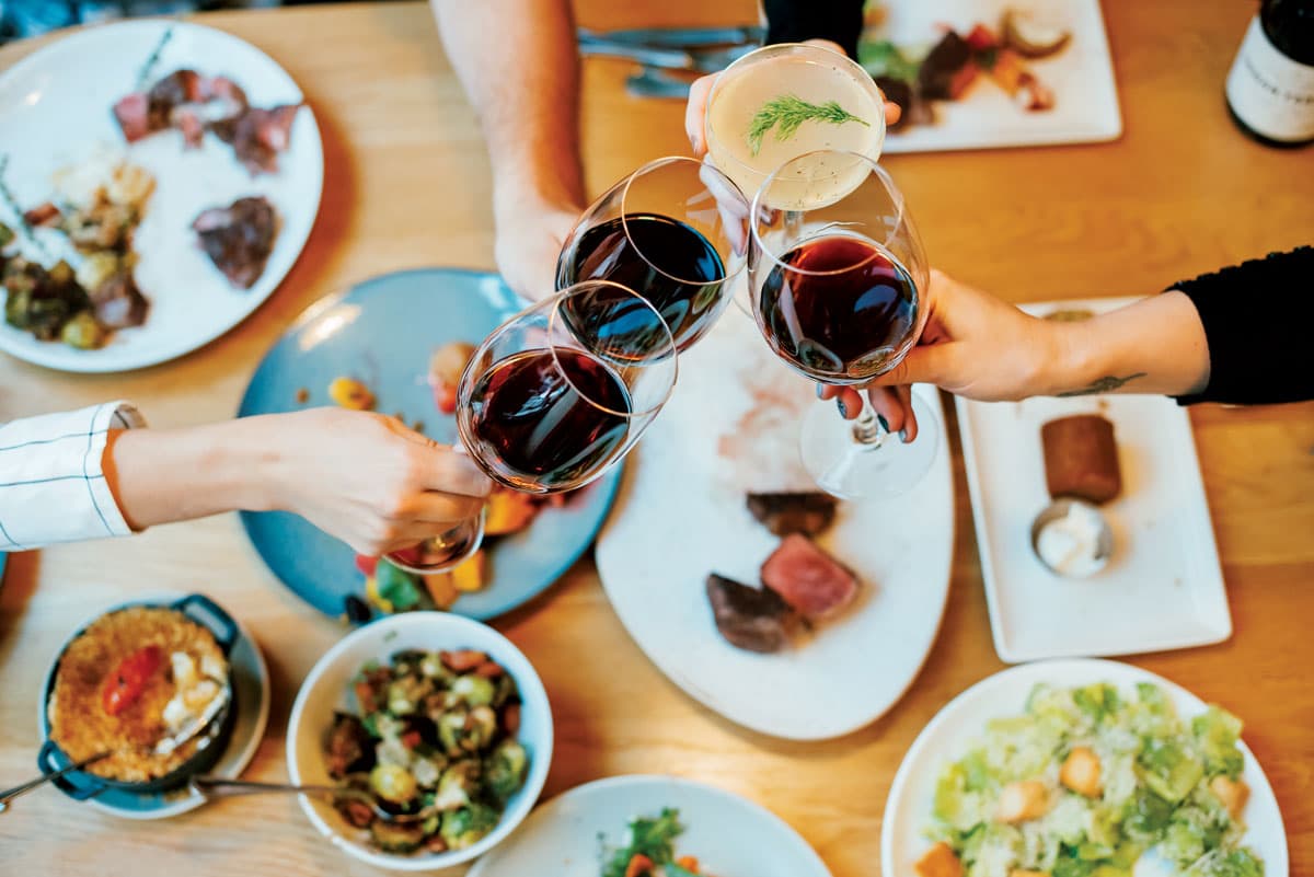 Three stemmed glasses of red wine clink over a table filled with plates and bowls of half-eaten delicacies at Urban Farmer in Denver, Colorado.