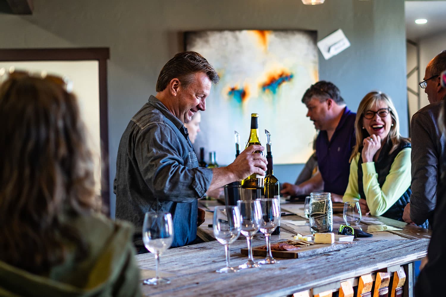 Smiling people gathered around a bar where a cheerful person is pouring wine into glasses from a green bottle in Palisade Colorado