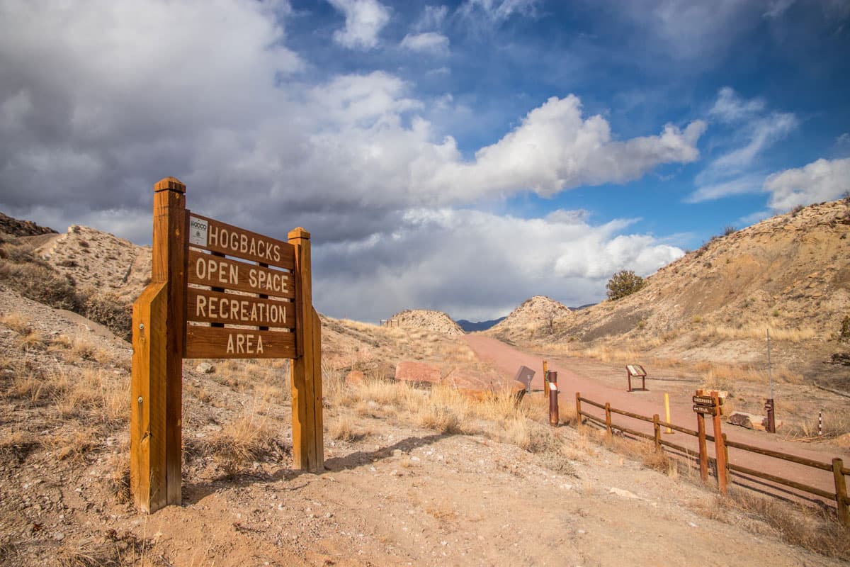 A wooden sign in the foreground labeling a trailhead that says "Hogbacks Open Space Recreation Area." In the background is a cloudy sky above a hiking trail path.