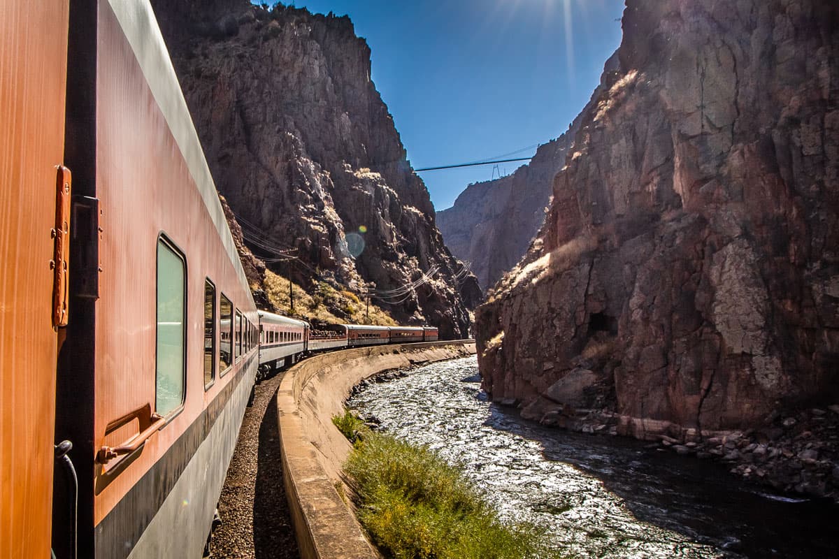 The red Royal Gorge train runs along on the railroad at the bottom of a wide valley. In the background is the Royal Gorge bridge against a deep blue sky.