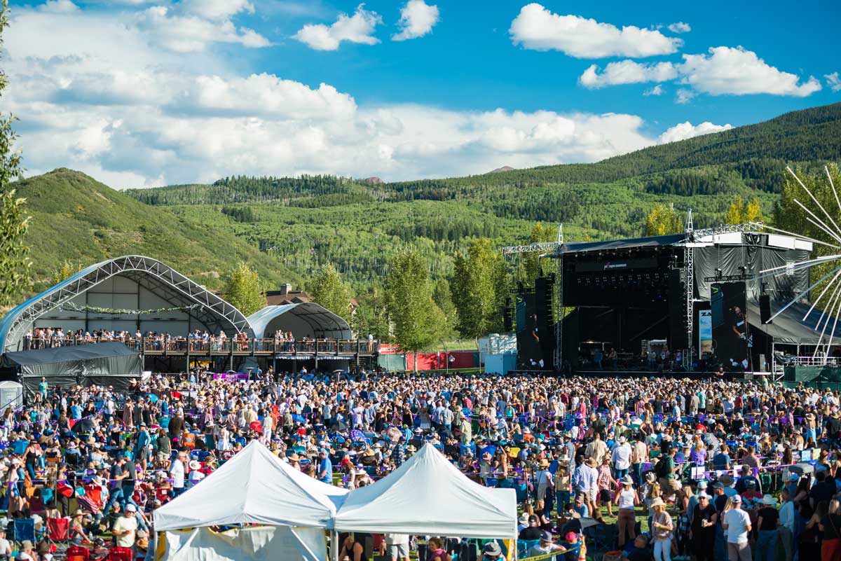 A crowd gathers at two stages with green mountains behind them to listen to bands at the JAS Labor Day Experience in Snowmass
