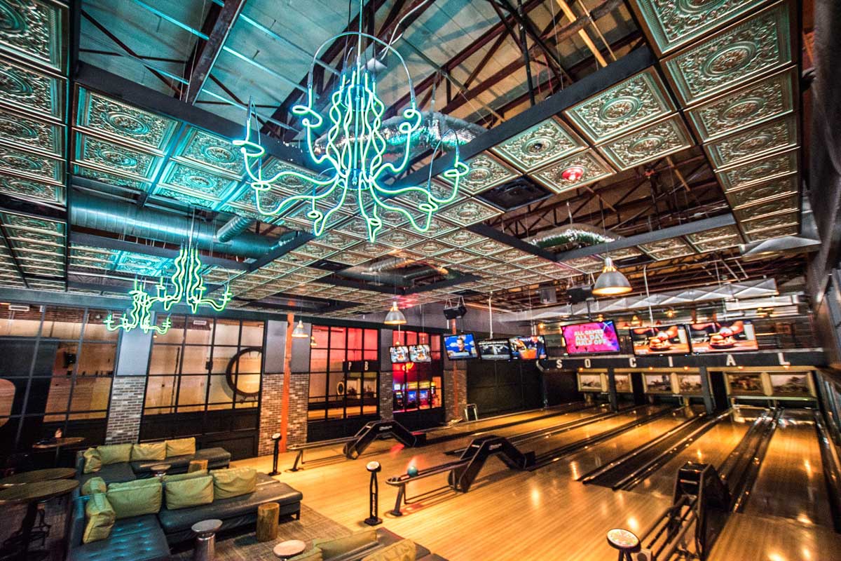 Neon-green chandeliers rise above a sleek bowling alley empty of patrons beneath a tin ceiling.