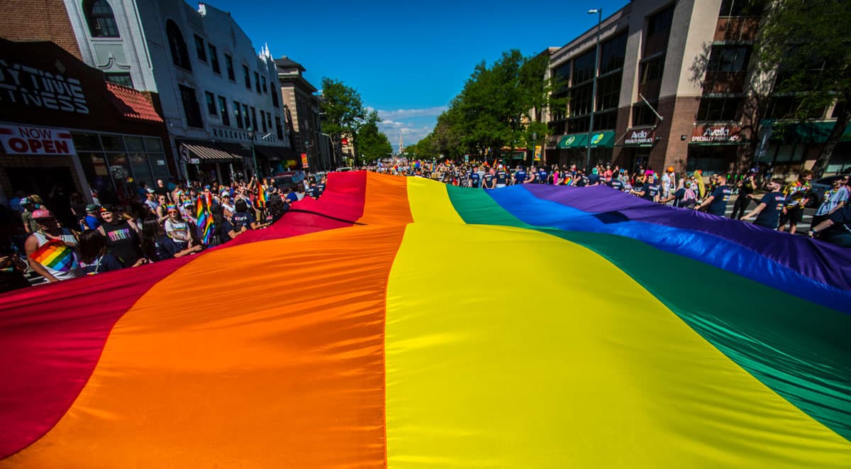 In the foreground is a close-up, wide shot of large a rainbow flag stretching across a street in Denver. There are lots of people with small rainbow flags dressed for summer.