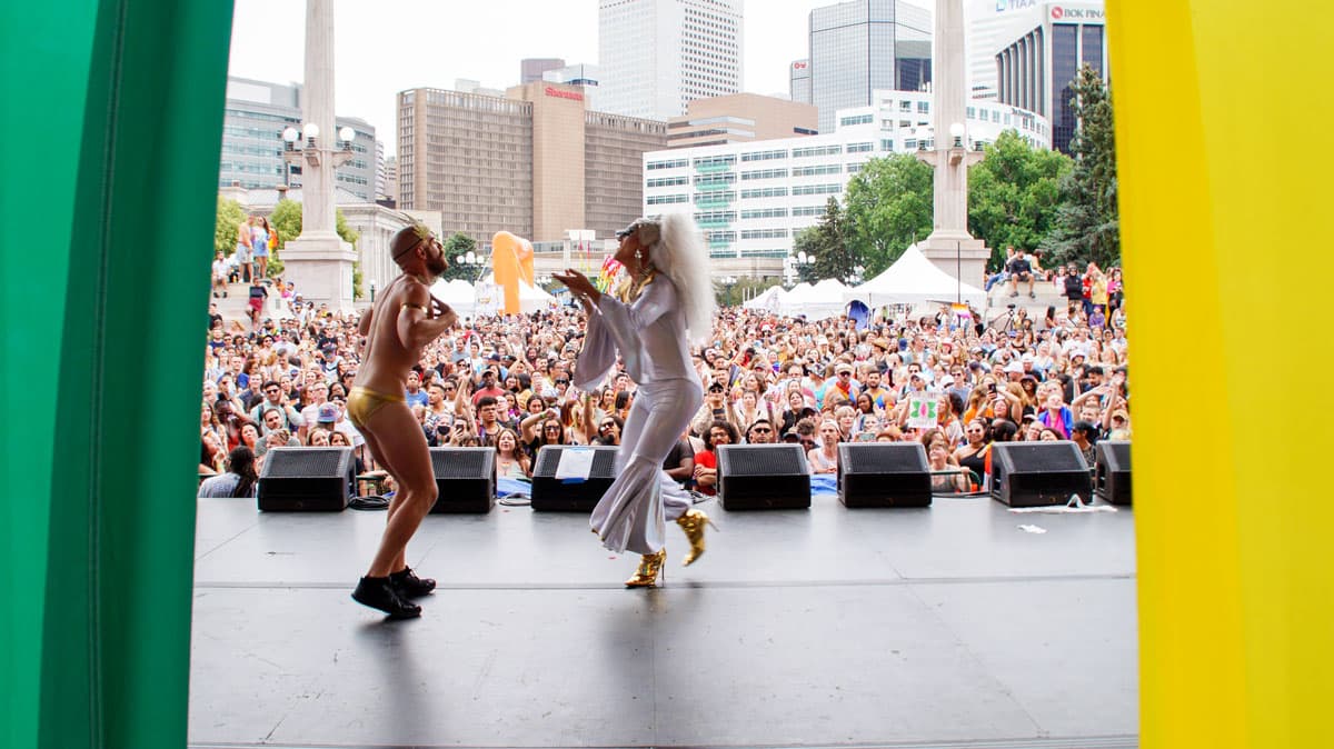 Two people perform on stage for a large audience. One is wearing tight, golden short and the other wears a white jumpsuit with flared arms and legs in Denver.
