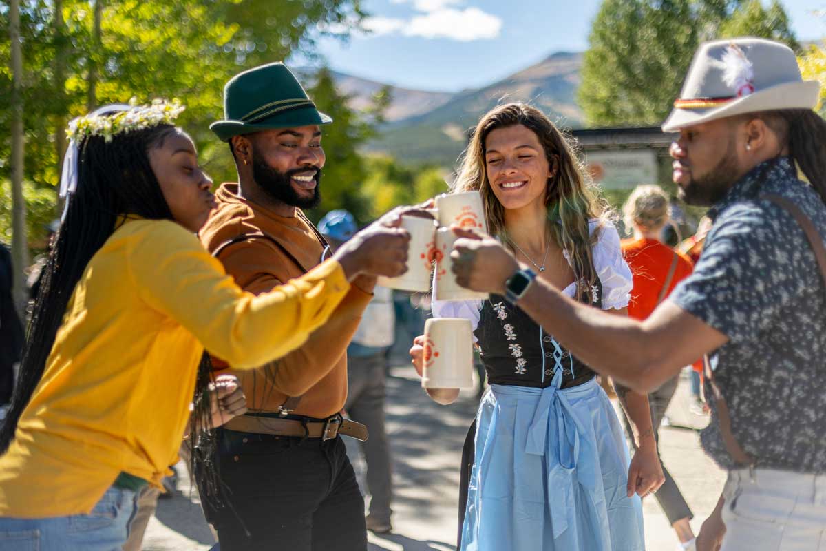 Four people in German-style dress clink and toast their beer steins under sunny skies at the Breckenridge Oktoberfest. In the distance, you can see shadowed mountain peak.