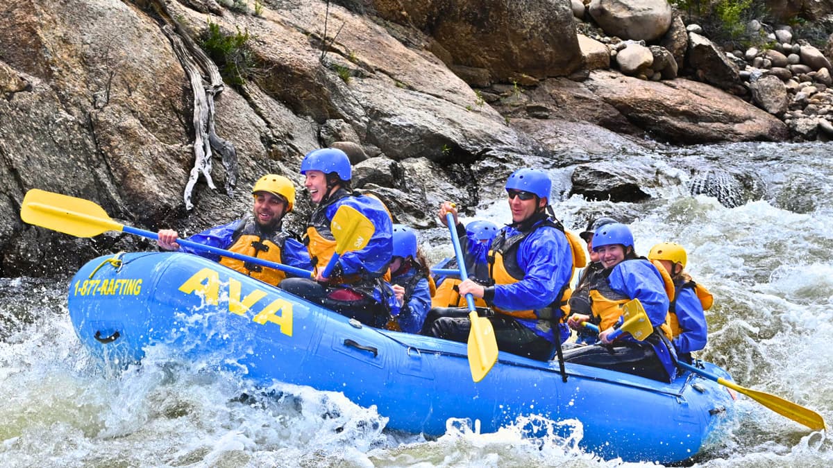 A group of about 10 people in protective gear and blue wetsuits sit on an inflated blue whitewater raft that says AVA on the side, as part of a whitewater rafting trip with Arkansas Valley Adventures Rafting & Zipline. The people on the raft are smiling and holding paddles, splashing in rollicking waves beside moss-covered rock.