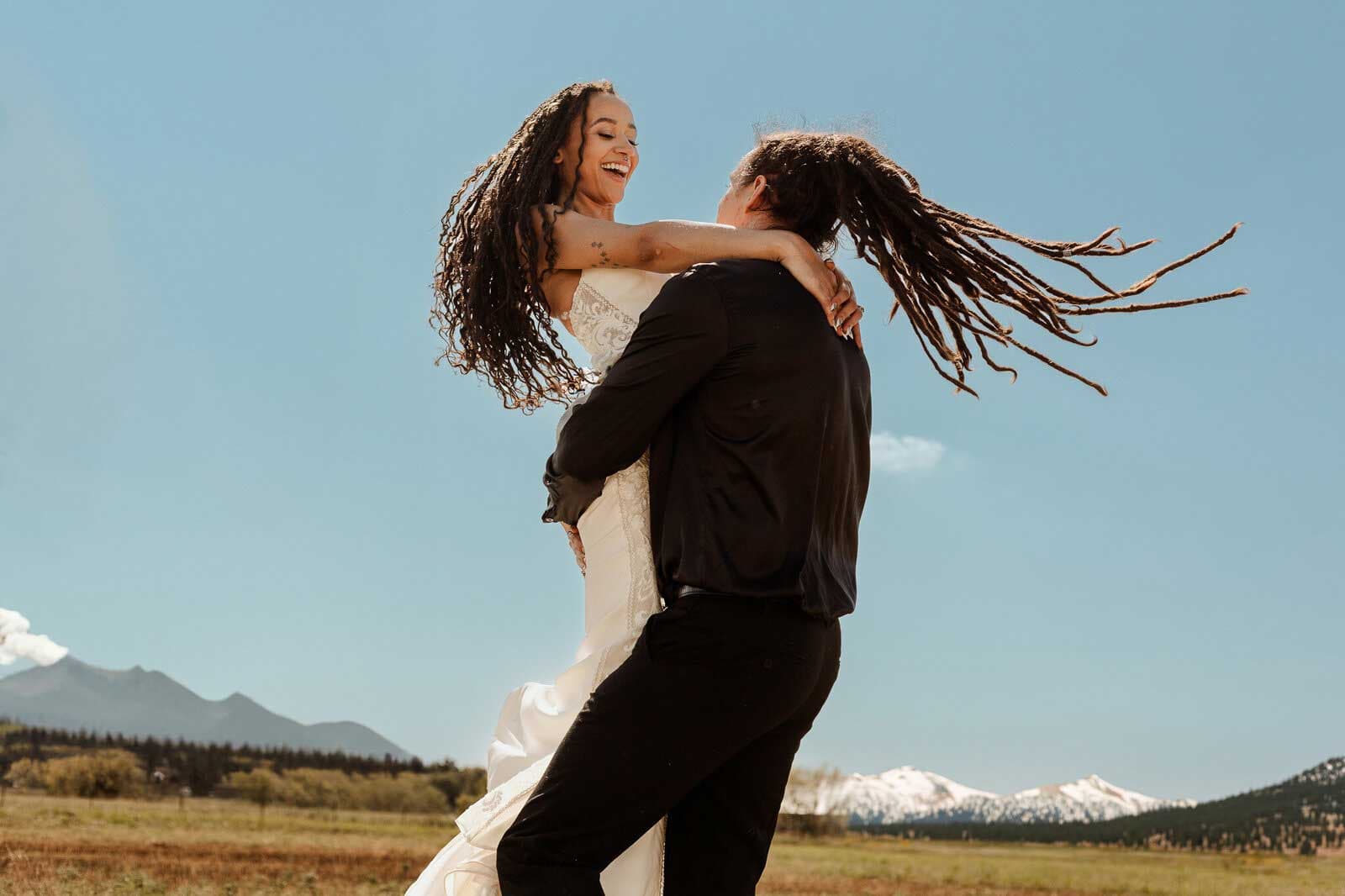 A couple in a black tux and white wedding gown twirl around under blue skies and mountains in the distance