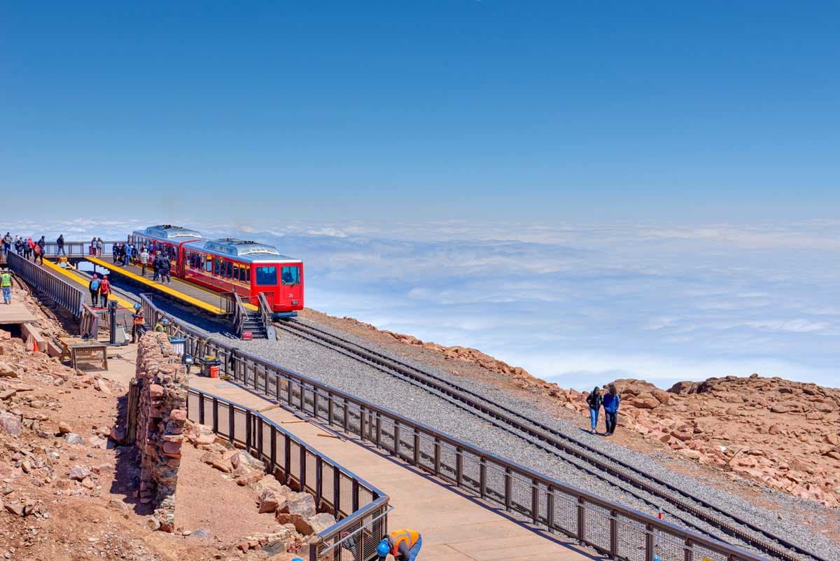A red train climbs a mountain peak where visitors wait at the summit