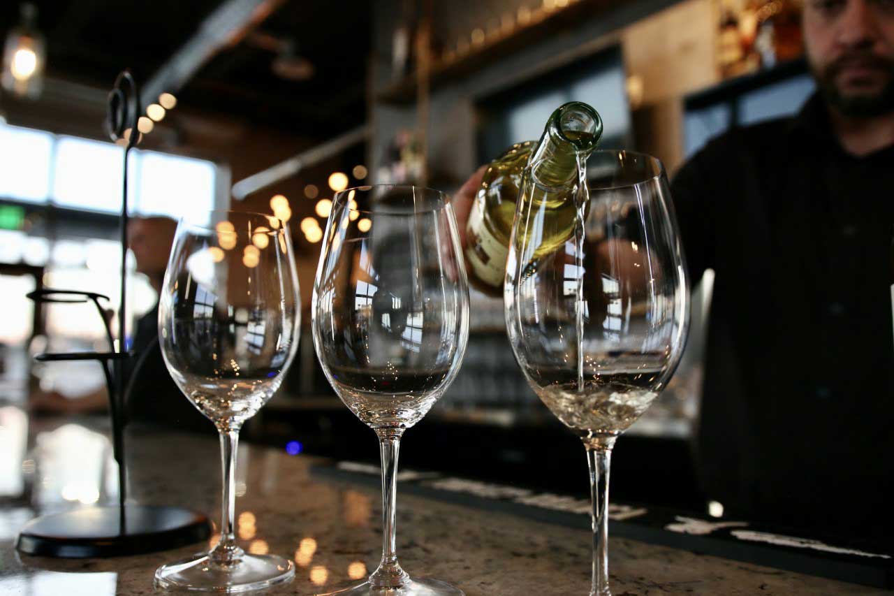 A bartender pours white wine into one of three glasses sitting on the bar