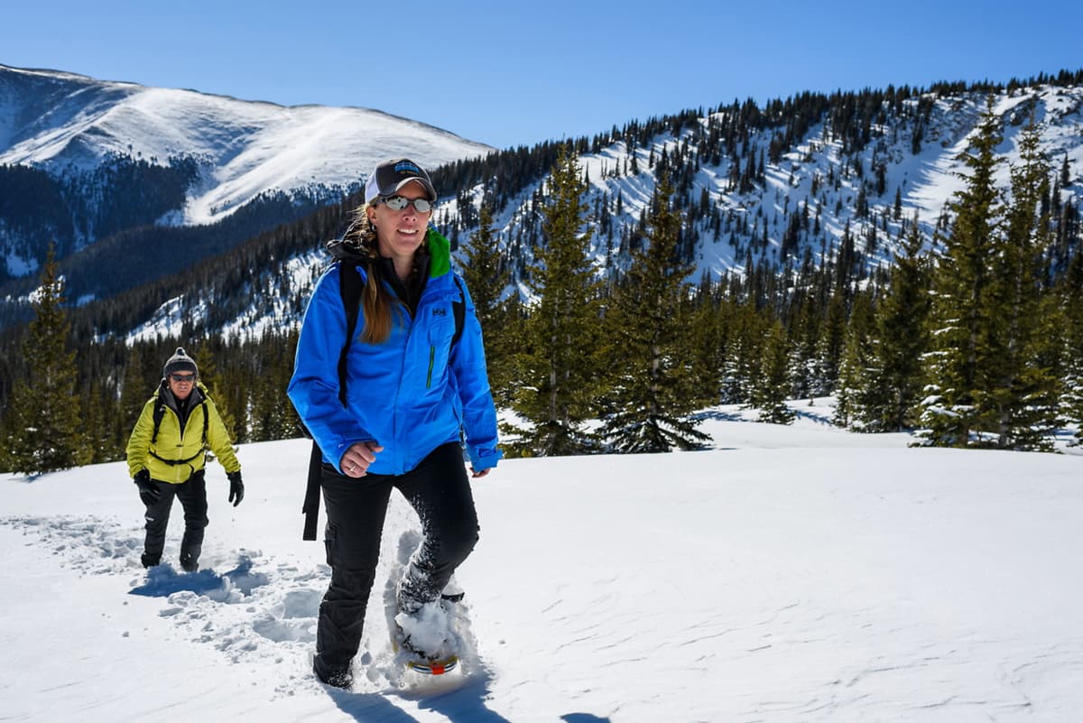 Two people dressed in warm clothes walk on snowshoes through a few inches of fresh powder in Winter Park, Colorado. All around them are hills peppered with pine trees, all under a clear blue sky.