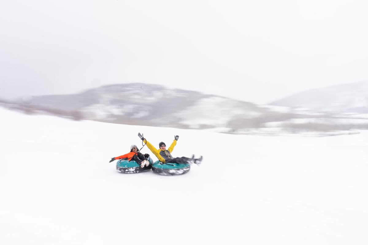 Two laughing and smiling people in their respective blue-and-black snow tubes tied to each other slide down a white, snowy hill as it snows outside. One of the people throws their hands up in the air looking at the camera, and the person behind them has their arms out wide beside them, looking at the person in front.