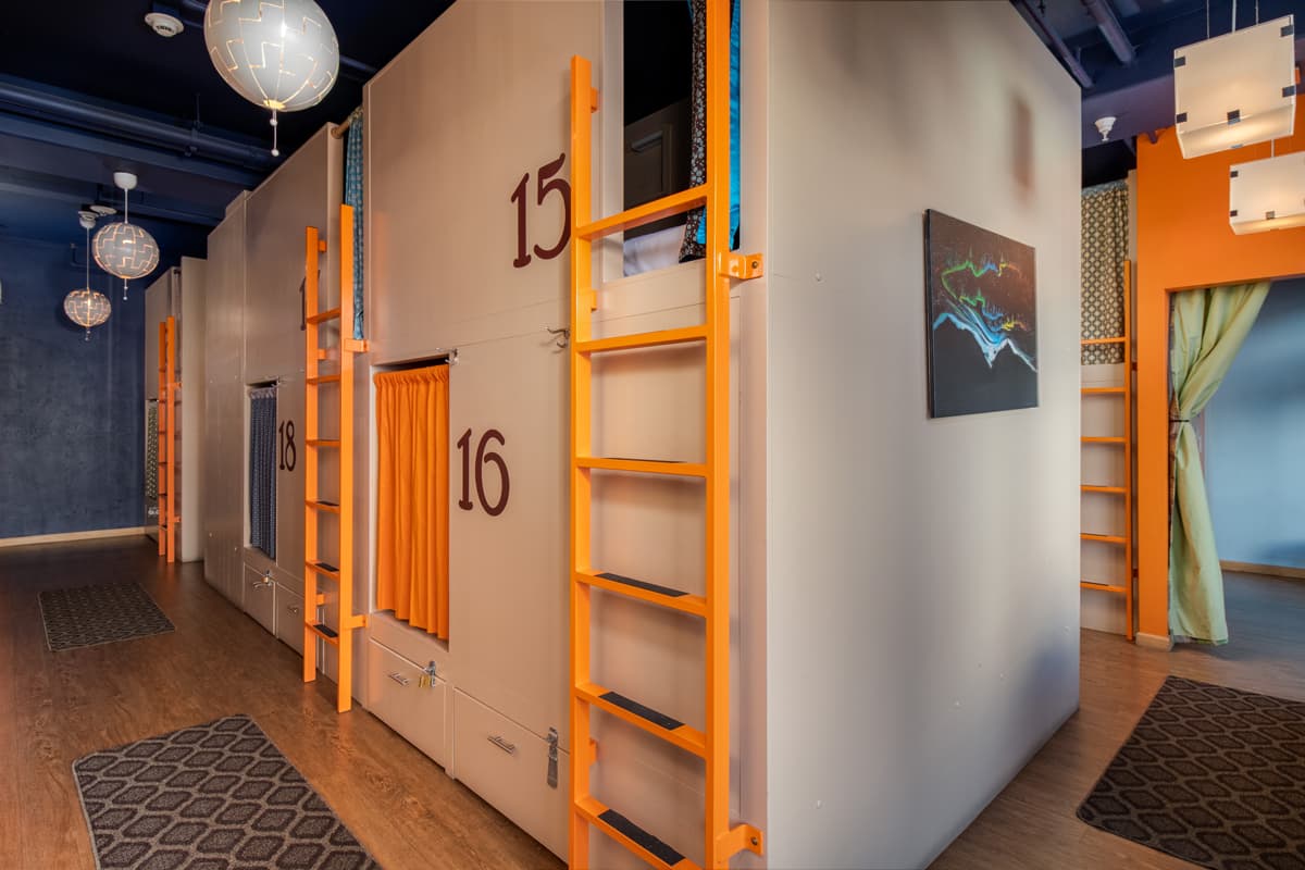 The interior of the Bunkhouse hostel in Minturn, Colorado. On the righthand side of the photo is a beige wall with six rectangular doorways within the wall, three of which have an orange ladder leading up to the doorway, covered in a dark blue curtain. Beyond the curtains are padded white-linen-covered mattresses. Along the sides of each doorway are painted numbers from 15 through 20. Hanging from the ceiling are several spherical lighting fixtures as well as two square ones.