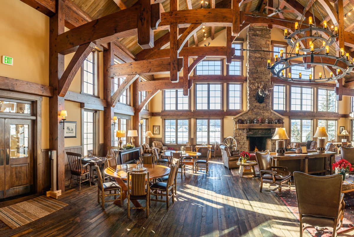 A tall-ceilinged, tall, elaborate hall decorated with wooden beams, lots of square windows, warm brown paint on the walls, a large circular chandelier hanging from the ceiling and a tall, broad stone fireplace. On the floor of the hall sit several wooden tables, cushiony seats, lamps, rugs and Wild West decorations.