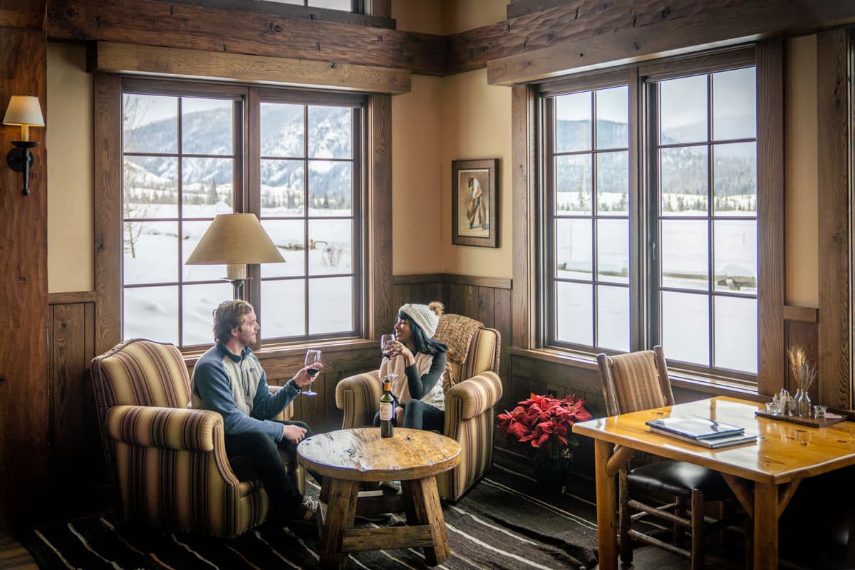 Two smiling and warmly dressed people have glasses of red wine in their hands, sitting in comfy chairs in a cozy corner of a dining hall at Vista Verde Guest Ranch in Clark, Colorado. The windows around them display the distant snow-covered mountains.