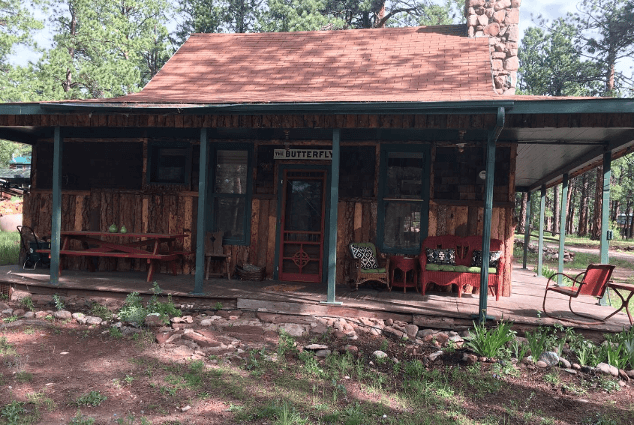 A cherry-colored picnic table and chairs stand under the porch of The Butterfly cabin. Trees stand tall around it.