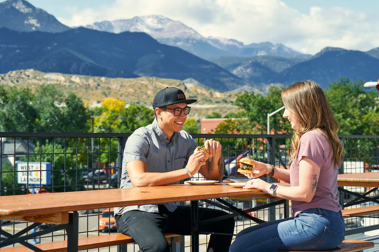 Two people sit at a rooftop table, eating lunch with mountains in the background
